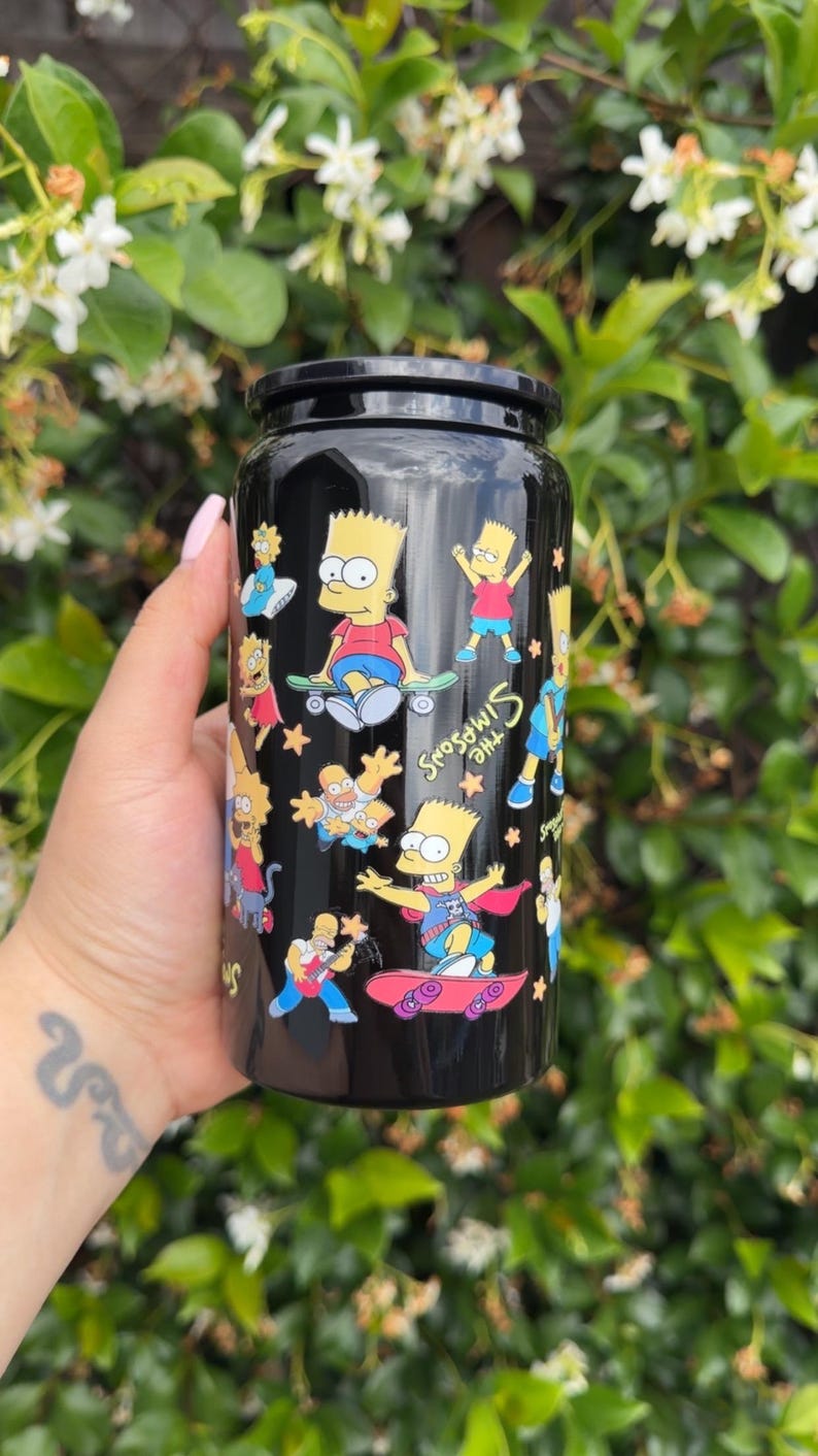 Pode incluir: Caneca preta com um design com personagens dos Simpsons. A caneca tem uma tampa preta e um acabamento brilhante. O design inclui Bart Simpson andando de skate e as palavras "The Simpsons".