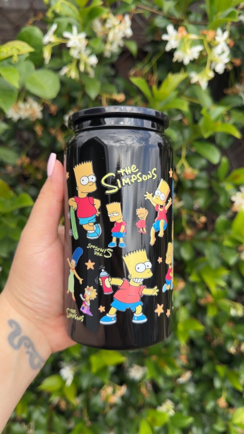 Pode incluir: Caneca preta com ilustra&ccedil;&otilde;es de Bart Simpson da s&eacute;rie de desenhos animados Os Simpsons. A caneca tem uma tampa preta e as palavras "The Simpsons" em amarelo. Bart &eacute; retratado em v&aacute;rias poses, com estrelas espalhadas pelo design.