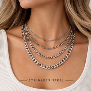 Puede incluir: Un conjunto de cuatro collares de cadena de acero inoxidable en tono plateado superpuestos. Los collares presentan eslabones de distintos tamaños, desde una cadena fina y delicada en la parte superior hasta una cadena gruesa en la inferior.