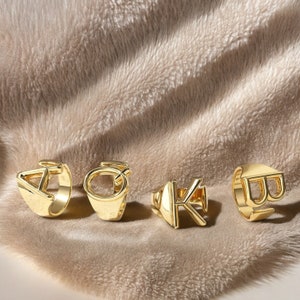 May include: Four gold-toned rings with different letters, A, O, K, and B, are displayed on a soft, fuzzy, light brown surface.