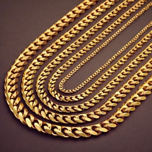 May include: A collection of gold-colored chain necklaces in varying thicknesses, arranged in a fan-like display. The chains have a classic curb link design, reflecting light and creating a luxurious appearance. The background is a dark, neutral color.