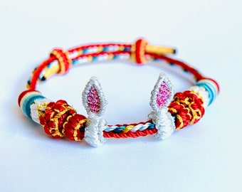 May include: A red, white, and blue braided bracelet with two white bunny ears with pink glitter accents.