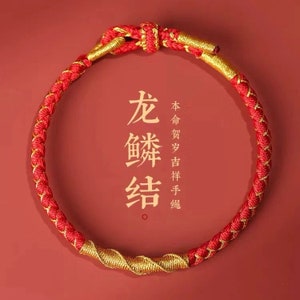 Könnte beinhalten: Ein rot-goldenes geflochtenes Armband mit einem Knoten oben. Das Armband trägt chinesische Schriftzeichen, die "Langes Leben und Glück" bedeuten.