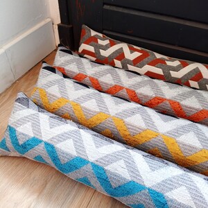 Peut inclure: Quatre coussins de porte avec un motif chevron dans des tons de gris, orange, jaune et bleu. Les coussins sont empilés les uns sur les autres.