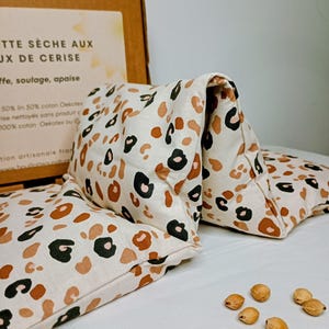 Peut inclure: Un coussin chauffant à imprimé léopard crème avec des taches marron, noires et beiges. Le coussin est plié et posé sur une surface blanche, avec plusieurs graines éparpillées à proximité. Une boîte en carton avec du texte français est en arrière-plan.