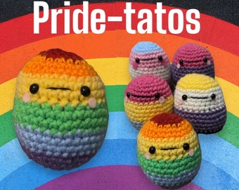 May include: A set of crocheted plushies in the shape of faces, each with a different color scheme representing the pride flag. The plushies are arranged on a rainbow background with the text "Pride-tatos" above them.