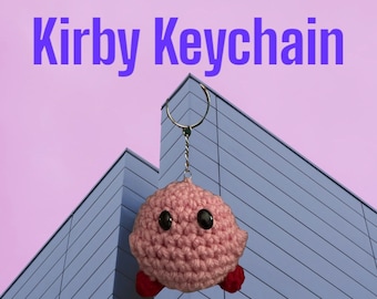 May include: A pink crocheted Kirby keychain hanging from a silver chain against a blue and gray background. The text "Kirby Keychain" is at the top of the image.