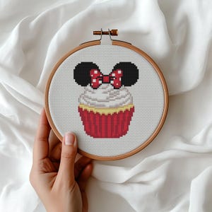 May include: A circular wooden embroidery hoop containing a cross-stitch pattern of a cupcake topped with black mouse ears and a red bow with white polka dots. The cupcake has white frosting and a red striped liner on a white fabric background.