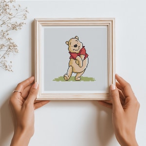 May include: A framed cross-stitch of Winnie the Pooh. The cartoon bear is depicted in a playful pose, wearing a red shirt. The background is a light gray, and the frame is a light wood color.