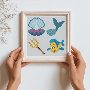May include: A square framed cross-stitch pattern featuring four distinct icons: an open clam shell with a pearl, a blue mermaid tail, a golden trident, and a yellow and blue tropical fish, all displayed on a light gray grid background.
