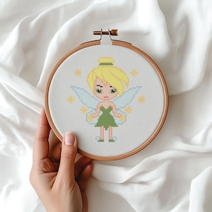 Puede incluir: Un bastidor de bordado de punto de cruz con una hada de dibujos animados con cabello rubio, ojos verdes y un vestido verde. El hada tiene alas de color azul claro y está rodeada de acentos amarillos en forma de estrella. El bordado está en un bastidor de madera.