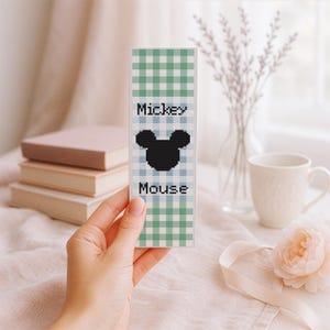 Op de afbeelding: Handgemaakte boekenlegger met een zwart Mickey Mouse silhouet op een blauw en groen geruit patroon. De tekst "Mickey Mouse" staat boven en onder het silhouet. De boekenlegger wordt in een hand gehouden.