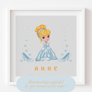 Peut inclure: Une broderie au point de croix encadrée représentant une princesse de dessin animé en robe bleue, avec des chaussures assorties de chaque côté. Le nom "ANNE" est brodé en lettres orange en dessous. Le texte est en bas.