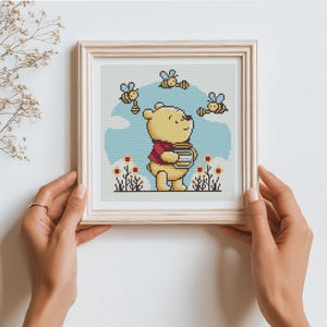 Puede incluir: Arte de punto de cruz de un oso amarillo con una camisa roja sosteniendo un tarro de miel, rodeado de abejas. El fondo presenta un cielo azul y flores. La obra de arte está enmarcada en un marco de madera clara.