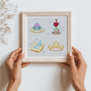 Peut inclure: Une &oelig;uvre d'art au point de croix encadr&eacute;e pr&eacute;sentant quatre motifs sur le th&egrave;me des contes de f&eacute;es. Les motifs comprennent un diad&egrave;me, une rose dans un d&ocirc;me en verre, un chausson de verre sur un coussin et une couronne. L'&oelig;uvre est dans un cadre en bois clair.