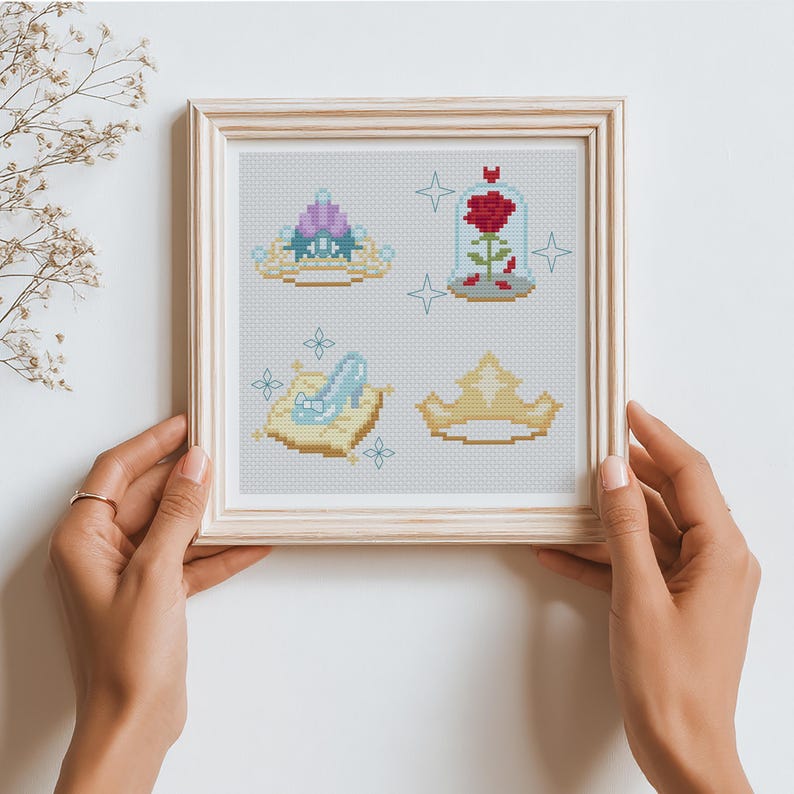 Peut inclure: Une &oelig;uvre d'art au point de croix encadr&eacute;e pr&eacute;sentant quatre motifs sur le th&egrave;me des contes de f&eacute;es. Les motifs comprennent un diad&egrave;me, une rose dans un d&ocirc;me en verre, un chausson de verre sur un coussin et une couronne. L'&oelig;uvre est dans un cadre en bois clair.