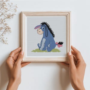 Könnte beinhalten: Kreuzstich-Kunst von Eeyore aus Winnie the Pooh, in hellem Holz gerahmt. Der Esel ist blau mit schwarzer Mähne, rosa Schleife und sitzt auf grünem Gras. Der Hintergrund ist hellgrau.