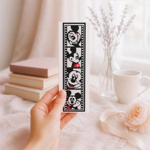 May include: A handmade bookmark featuring a black and white cross-stitch design of Mickey Mouse in four different poses. The bookmark has a white border and red accents. The background includes books, a cup, and a flower.