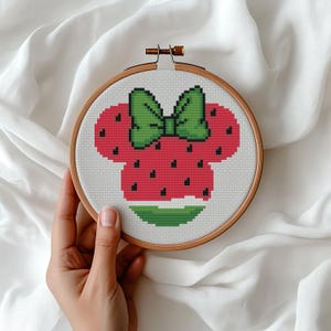 Op de afbeelding: Een borduurring met kruissteek met een aardbei-ontwerp met een groene strik. De aardbei is rood met zwarte zaden en de strik is groen. De borduursel zit in een houten ring.