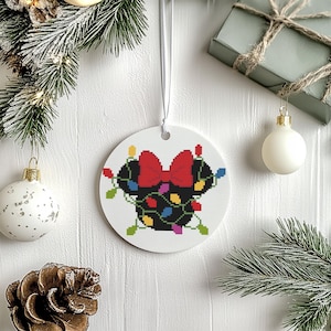 May include: A white, circular Christmas decoration featuring a black mouse head silhouette with a red bow and colourful fairy lights. The ornament hangs from a white ribbon. The background includes festive decorations.