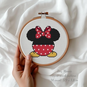 May include: A cross-stitch pattern of Minnie Mouse in a wooden embroidery hoop. The design features Minnie's head, bow, and dress in black, red, and white. The text "Tiny Minnie cross stitch pattern" is below the design. The pattern is 3.9 x 3.8 inches.