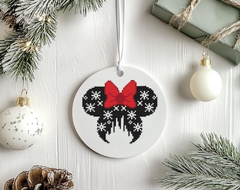 May include: A white ceramic Christmas ornament featuring a black and white pixelated design of a character's silhouette with a red bow. The ornament is hanging from a white ribbon, with holiday decorations in the background.