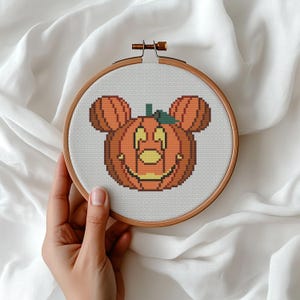 May include: A circular wooden embroidery hoop containing white Aida fabric with a cross-stitch design of an orange pumpkin shaped like a mouse head. The pumpkin features a green stem, yellow eyes, a yellow nose, and a wide yellow stitched mouth.