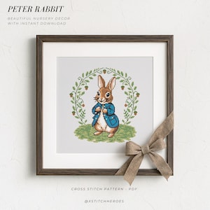 May include: A square wooden frame containing a cross-stitch pattern of Peter Rabbit wearing a blue jacket, standing on green grass within an acorn and leaf wreath. Text reads PETER RABBIT, BEAUTIFUL NURSERY DECOR WITH INSTANT DOWNLOAD, CROSS STITCH PATTERN - PDF, @XSTITCHHEROES.