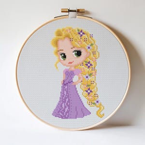 May include: A cross-stitch embroidery of a cartoon princess with long blonde hair decorated with purple flowers, wearing a purple dress. The artwork is displayed in a wooden embroidery hoop, set against a white backdrop.