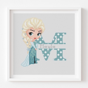 May include: A cross-stitch pattern featuring a cartoon character with blonde hair in a braid, wearing a blue dress, positioned next to a large letter M in blue with white polka dots and the name Mayla written in the centre of the letter.