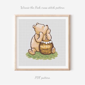 May include: A cross stitch pattern featuring a light tan bear sitting on a patch of green grass, leaning against a brown honey pot with the word HUNNY printed on it. The design is displayed within a light wood frame against a white background.
