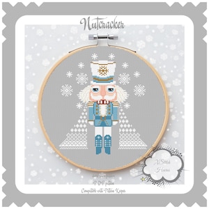 Puede incluir: Patrón de punto de cruz de un Cascanueces azul y blanco con bigote rojo y sombrero blanco. El Cascanueces está de pie frente a dos árboles de Navidad blancos. El patrón está sobre un fondo gris con copos de nieve blancos. El texto "X Stitch Heaven" está en la esquina inferior derecha de la imagen. El texto "Cross Stitch Pattern - PDF Pattern - Compatible with Pattern Keeper" está en la esquina inferior izquierda de la imagen.