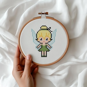 Puede incluir: Un bastidor de bordado de punto de cruz con un diseño de hada pixelada. El hada tiene cabello rubio, alas azules y un vestido verde. El bordado está en un bastidor de madera, ideal para la decoración del hogar o proyectos de manualidades.
