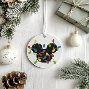 Peut inclure: Ornement de Noël rond et blanc avec une silhouette noire de tête de souris ornée de guirlandes lumineuses colorées. L'ornement est suspendu à un ruban blanc. D'autres décorations de Noël sont en arrière-plan.