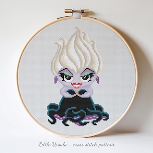 Pode incluir: Um padrão de ponto cruz de Ursula de A Pequena Sereia, exibido em um bastidor de bordado de madeira. A personagem de desenho animado tem cabelo branco, pele roxa e um vestido preto. O texto "Little Ursula - cross stitch pattern" está na parte inferior.