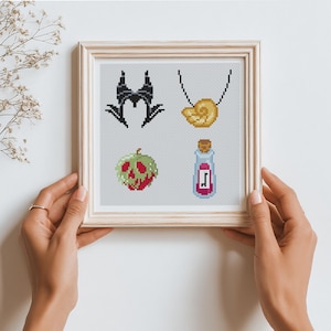 May include: Cross-stitch art in a wooden frame featuring four designs: a black horned headdress, a gold shell necklace, a green and red poisoned apple, and a potion bottle with a cork. The artwork is held by two hands.