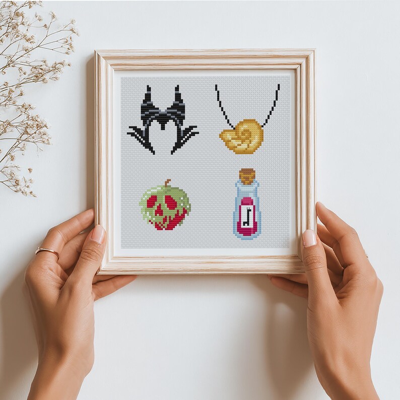 May include: Cross-stitch art in a wooden frame featuring four designs: a black horned headdress, a gold shell necklace, a green and red poisoned apple, and a potion bottle with a cork. The artwork is held by two hands.