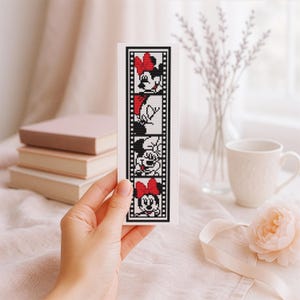 Puede incluir: Un marcapáginas blanco con un diseño de punto de cruz de Minnie Mouse en un estilo de tira de película. El diseño en blanco y negro incluye la cara de Minnie y un lazo rojo. El marcapáginas se sostiene frente a un fondo suave y neutro.