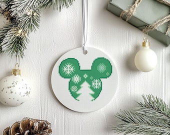 May include: A white, round Christmas ornament with a green pixelated design of a mouse head and a Christmas tree. The ornament is hanging by a white ribbon. Other Christmas decorations are in the background.