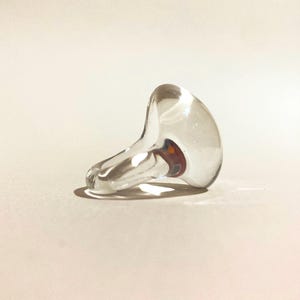 Puede incluir: Un tapón de botella de vidrio transparente con una parte superior redondeada y una base acampanada. El tapón tiene un pequeño detalle interior de color naranja oscuro. El objeto descansa sobre una superficie beige claro.