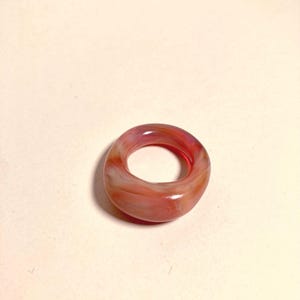 May include: A translucent ring with a marbled appearance. The ring features shades of red, pink, and white, creating a swirled effect. The ring is a solid band with a smooth, polished surface. Jewelry.