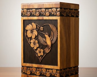 May include: Wooden urn with a carved hummingbird and hibiscus design. The rectangular box features a heart-shaped panel with a hummingbird in flight, surrounded by flowers. The top and bottom edges have a carved leaf pattern.
