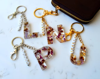 Op de afbeelding: Vier sleutelhangers van hars in de vorm van letters met gedroogde bloemen en gouden vlokken. Elke sleutelhanger heeft een zilveren of gouden ketting en een zilveren of gouden karabijnhaak. De letters zijn L, N, P en J. De namen die op de sleutelhangers zijn gespeld, zijn Laura, Nadia, Perla en Jessica.