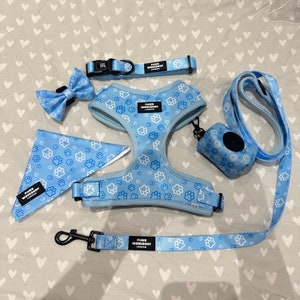 May include: A light blue dog accessory set including a collar, harness, lead, bow tie, bandana, and waste bag dispenser. Each item is patterned with white paw prints. The brand name "PAWS WORKSHOP LONDON" is printed on each item.