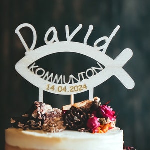 May include: A layered cake topped with a wooden cake topper. The topper is shaped like a fish and has the name "David", the word "Kommunion", and the date "14.04.2024" cut out. The cake is decorated with flowers and other decorative elements.