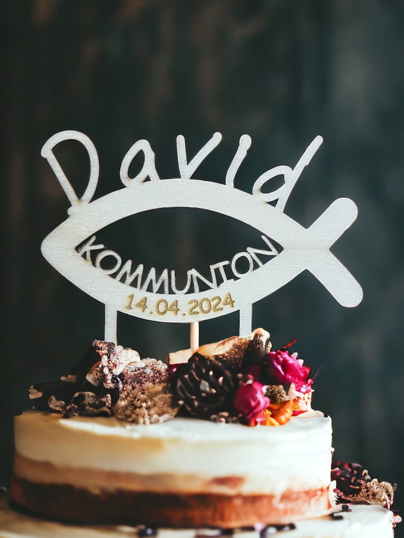 May include: A layered cake topped with a wooden cake topper. The topper is shaped like a fish and has the name "David", the word "Kommunion", and the date "14.04.2024" cut out. The cake is decorated with flowers and other decorative elements.