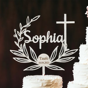 May include: A white tiered cake with red roses and a decorative topper. The topper features the name "Sophia" in script, a cross, and the text "Taufe 16.04.2021". A small figurine of a couple is on top.