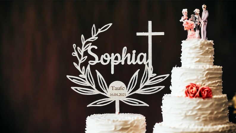 May include: A white tiered cake with red roses and a decorative topper. The topper features the name "Sophia" in script, a cross, and the text "Taufe 16.04.2021". A small figurine of a couple is on top.
