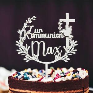May include: A white cake topper with the words "Zur Kommunion Max" in script, featuring a cross and dove design. The topper is placed on a chocolate cake decorated with colorful candies and frosting. The cake is a centerpiece for a religious celebration.