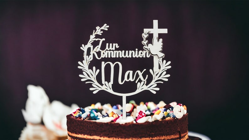 May include: A white cake topper with the words "Zur Kommunion Max" in script, featuring a cross and dove design. The topper is placed on a chocolate cake decorated with colorful candies and frosting. The cake is a centerpiece for a religious celebration.